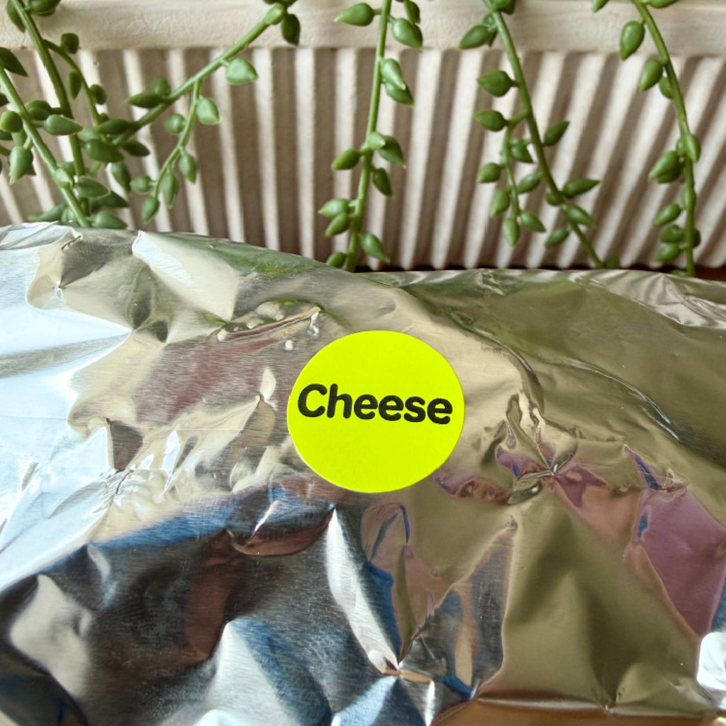 A crumpled silver foil package with a bright yellow circular sticker that reads 'Cheese' in black text, photographed close up with trailing green plants and a slatted cream background behind it.