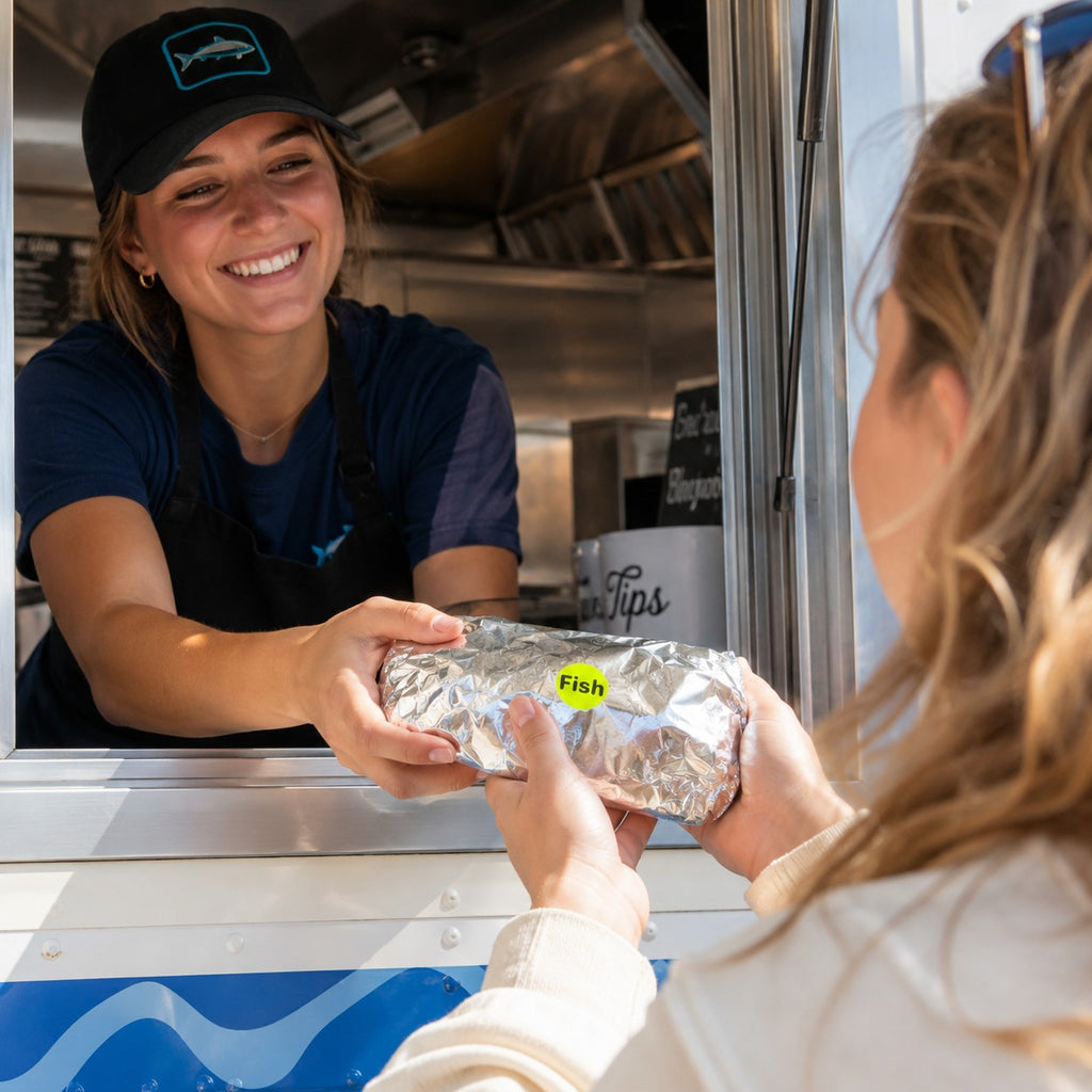 A smiling food vendor in a navy uniform and black cap hands a foil-wrapped food item to a customer at a food truck window. The vendor wears a cap with a blue fish logo, and a small yellow 'Fish' sticker is visible on the food wrap.