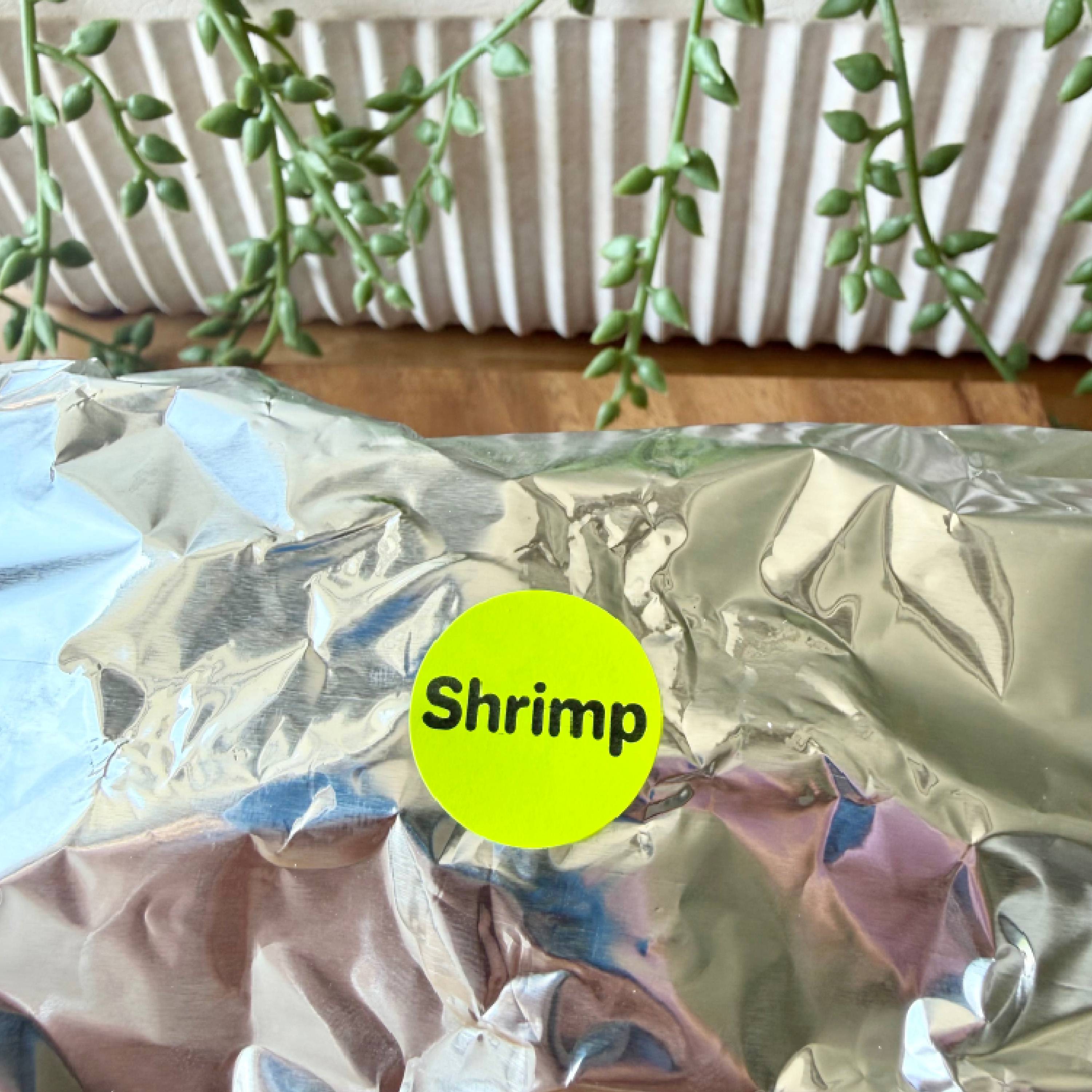A crinkled silver foil package with a bright yellow circular sticker reading 'Shrimp' in black text, placed on a wooden surface with green trailing plants and a white radiator in the background.